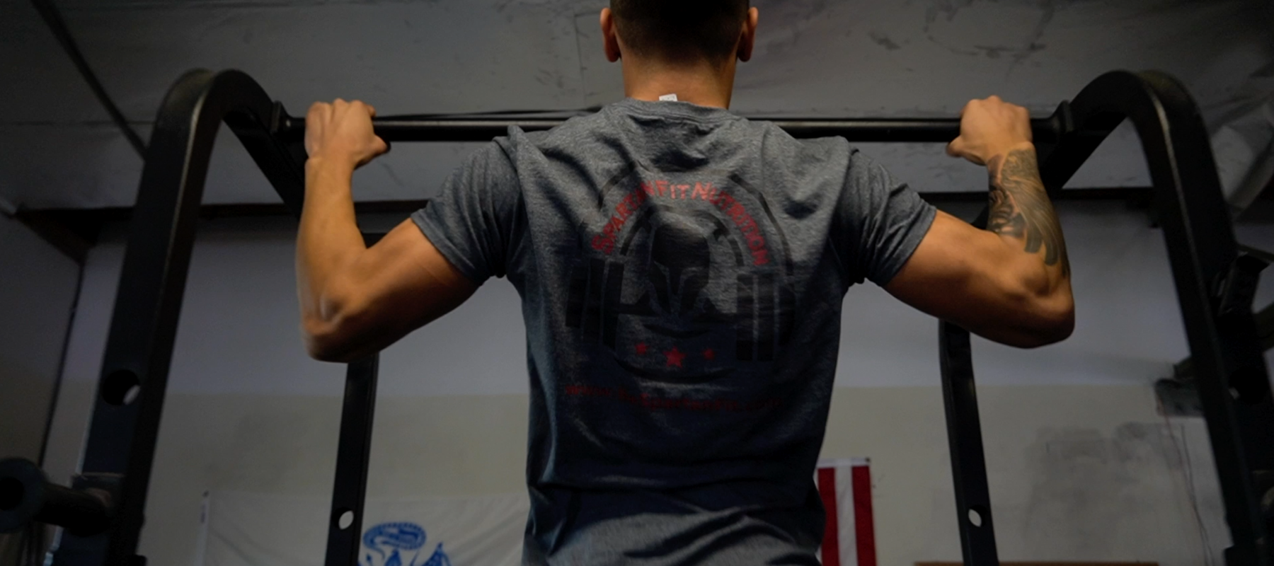 Bodybuilding athlete wearing Spartan Fit Nutrition T-Shirt doing pull up exercise at Fitness Gym.